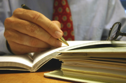 a hand holding a pencil, writing in a notebook, next to glasses and a stack of papers and books